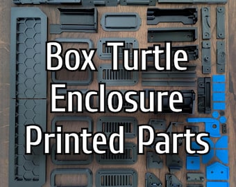 Op de afbeelding: Een assortiment donkergrijze en blauwe 3D-geprinte onderdelen voor een Box Turtle Enclosure, gerangschikt op een houten oppervlak. De onderdelen omvatten verschillende vormen en maten, zoals rechthoekige frames, ventilatieopeningen en panelen. De tekst "Box Turtle Enclosure Printed Parts" is zichtbaar.