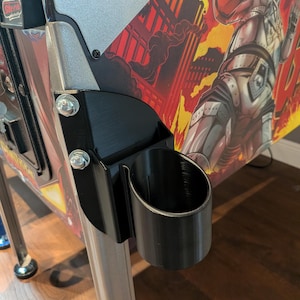 May include: A black plastic cup holder attached to a pinball machine. The holder is mounted with silver bolts and is positioned near the machine's side. The pinball machine has a colorful graphic design with a robot.