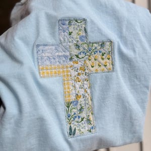 May include: Light blue sweatshirt with a quilted cross design on the back. The cross is made of fabric patches with floral and gingham patterns in yellow, blue, and white. The sweatshirt has a crew neck.