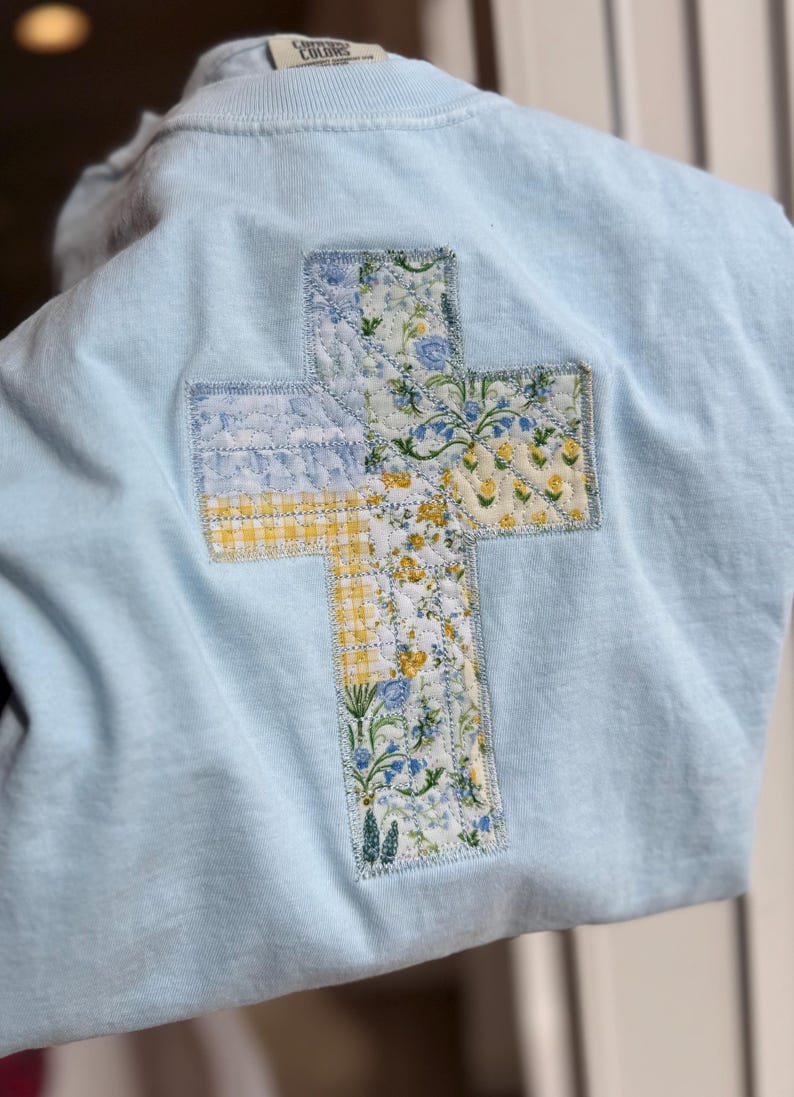 May include: Light blue sweatshirt with a quilted cross design on the back. The cross is made of fabric patches with floral and gingham patterns in yellow, blue, and white. The sweatshirt has a crew neck.