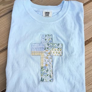 May include: A light blue Comfort Colors t-shirt with a floral cross applique. The cross features a patchwork design with yellow, blue, and white floral patterns. The shirt is folded on a wooden surface.