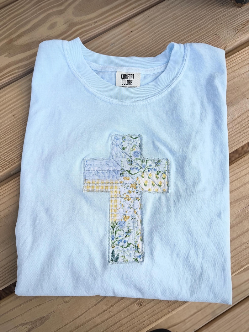 May include: A light blue Comfort Colors t-shirt with a floral cross applique. The cross features a patchwork design with yellow, blue, and white floral patterns. The shirt is folded on a wooden surface.