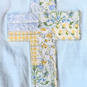 May include: A light blue fabric with a quilted cross applique. The cross is made of floral patterned fabric in yellow, blue, and white. The cross is stitched onto the light blue fabric with a light blue thread.