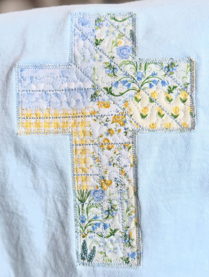 May include: A light blue fabric with a quilted cross applique. The cross is made of floral patterned fabric in yellow, blue, and white. The cross is stitched onto the light blue fabric with a light blue thread.