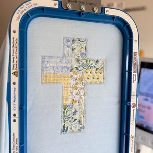 May include: A cross-shaped embroidery design in a blue embroidery hoop. The cross is composed of fabric patches with floral and gingham patterns in yellow, blue, and white. The hoop is blue and has text that reads "8"x13" Mighty Hoop".