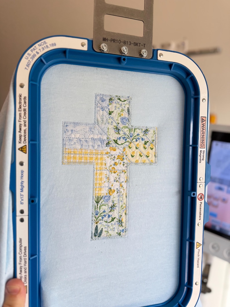 May include: A cross-shaped embroidery design in a blue embroidery hoop. The cross is composed of fabric patches with floral and gingham patterns in yellow, blue, and white. The hoop is blue and has text that reads "8"x13" Mighty Hoop".