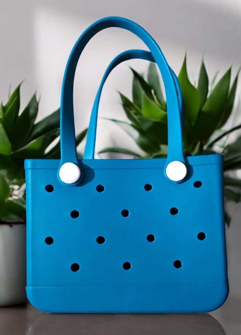 May include: A bright blue tote bag with two curved handles and a rectangular body. The bag features a series of evenly spaced, circular cutouts and white circular accents where the handles attach. The bag is made of a durable material, perfect for carrying items.
