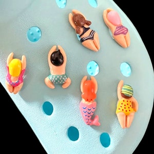 May include: A set of colorful, cartoon-style, plastic charms featuring swimmers and a mermaid. The charms are designed to be attached to shoes or other accessories.