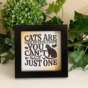 May include: A framed square sign with the text "CATS ARE LIKE POTATO CHIPS YOU CAN'T HAVE JUST ONE" in black lettering. A black cat silhouette and two hearts are also featured. The frame is black.