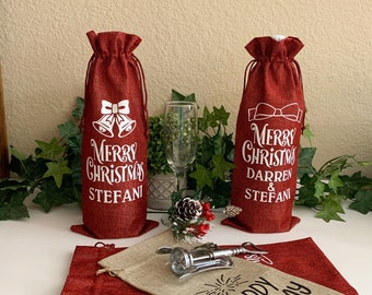 May include: Red wine bottle bags with white text that reads "Merry Christmas" and names. One bag has bells, the other a bow. A champagne flute, corkscrew, and other bags are also visible. Festive holiday gift.