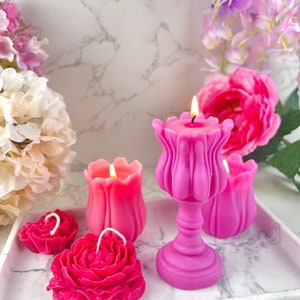 Pode incluir: Uma cole&ccedil;&atilde;o de velas rosas em v&aacute;rias formas florais, incluindo rosas e tulipas, com pavios acesos. As velas est&atilde;o dispostas numa bandeja com padr&atilde;o de m&aacute;rmore, rodeadas por flores rosa e roxas, criando um ambiente decorativo e rom&acirc;ntico.