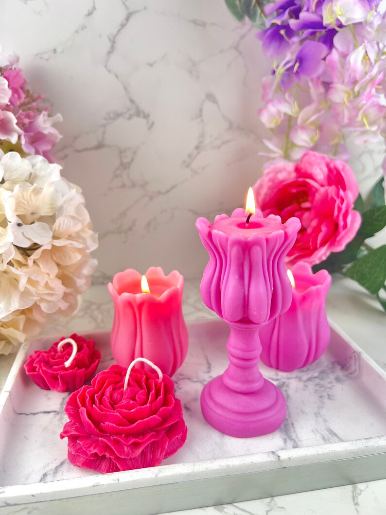 Pode incluir: Uma cole&ccedil;&atilde;o de velas rosas em v&aacute;rias formas florais, incluindo rosas e tulipas, com pavios acesos. As velas est&atilde;o dispostas numa bandeja com padr&atilde;o de m&aacute;rmore, rodeadas por flores rosa e roxas, criando um ambiente decorativo e rom&acirc;ntico.