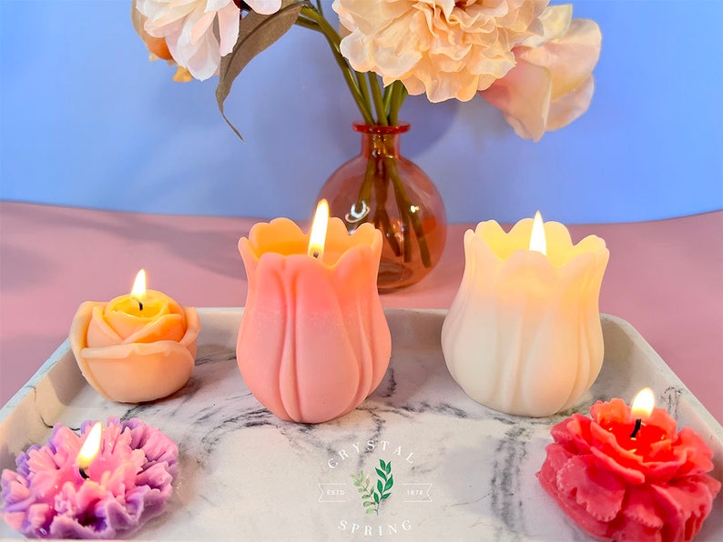 Pode incluir: Uma cole&ccedil;&atilde;o de velas acesas em forma de flor em v&aacute;rias cores, incluindo rosa, laranja e roxo, dispostas em uma bandeja de m&aacute;rmore. Um vaso de flores est&aacute; ao fundo. As velas t&ecirc;m a forma de rosa, tulipa e outros desenhos florais. A marca "CRYSTAL SPRING" &eacute; vis&iacute;vel.