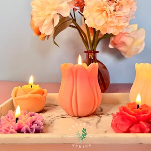 Pode incluir: Velas em forma de flor em v&aacute;rias cores, incluindo rosa, amarelo e vermelho, com pavios acesos. As velas est&atilde;o dispostas numa bandeja branca com design de m&aacute;rmore, juntamente com um vaso de flores cor de p&ecirc;ssego. O log&oacute;tipo Crystal Spring &eacute; vis&iacute;vel.