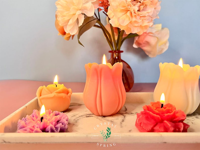 Pode incluir: Velas em forma de flor em v&aacute;rias cores, incluindo rosa, amarelo e vermelho, com pavios acesos. As velas est&atilde;o dispostas numa bandeja branca com design de m&aacute;rmore, juntamente com um vaso de flores cor de p&ecirc;ssego. O log&oacute;tipo Crystal Spring &eacute; vis&iacute;vel.