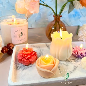 Pode incluir: Uma cole&ccedil;&atilde;o de velas acesas em v&aacute;rios formatos florais, incluindo uma rosa, tulipa e pe&ocirc;nia, exibidas em uma bandeja de m&aacute;rmore. Uma vela grande de vidro com o texto "Crystal Spring" tamb&eacute;m est&aacute; presente. A cena &eacute; decorada com flores.