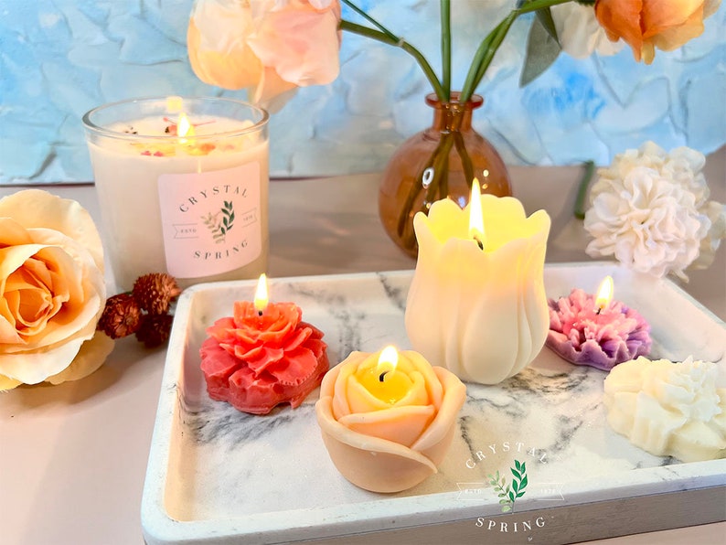 Pode incluir: Uma cole&ccedil;&atilde;o de velas acesas em v&aacute;rios formatos florais, incluindo uma rosa, tulipa e pe&ocirc;nia, exibidas em uma bandeja de m&aacute;rmore. Uma vela grande de vidro com o texto "Crystal Spring" tamb&eacute;m est&aacute; presente. A cena &eacute; decorada com flores.