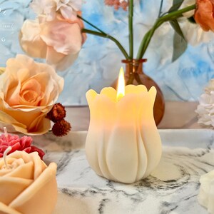 Pode incluir: Uma vela em forma de tulipa, de cor marfim, acesa e brilhante. A vela est&aacute; numa bandeja de m&aacute;rmore com outras velas florais. O fundo inclui rosas e um vaso com flores. O texto "CRYSTAL SPRING ESTD 1978" &eacute; vis&iacute;vel.