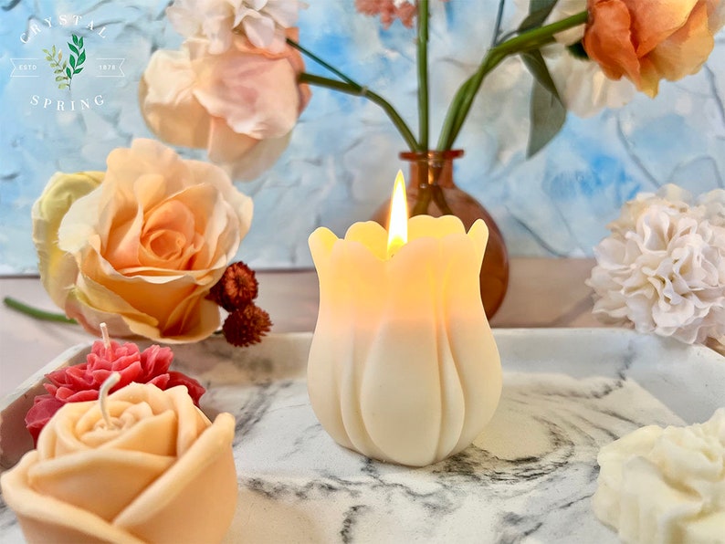 Pode incluir: Uma vela em forma de tulipa, de cor marfim, acesa e brilhante. A vela est&aacute; numa bandeja de m&aacute;rmore com outras velas florais. O fundo inclui rosas e um vaso com flores. O texto "CRYSTAL SPRING ESTD 1978" &eacute; vis&iacute;vel.