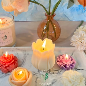 Pode incluir: Uma cole&ccedil;&atilde;o de velas acesas em v&aacute;rias formas florais, incluindo uma rosa, tulipa e cravo, exibidas em uma bandeja de m&aacute;rmore. Uma vela grande de vidro com o texto "Crystal Spring" tamb&eacute;m est&aacute; presente. A cena &eacute; complementada por um vaso de flores.