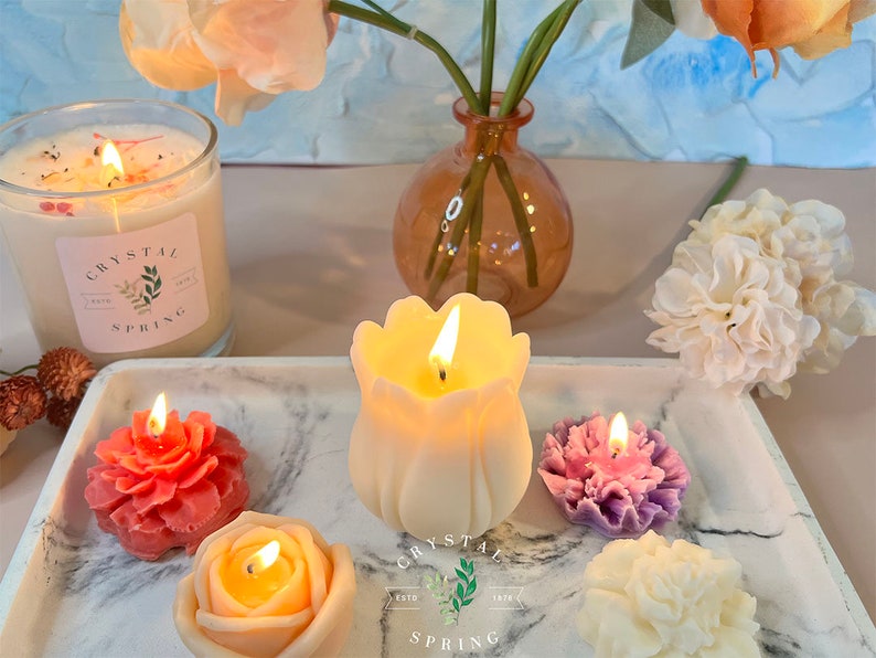 Pode incluir: Uma cole&ccedil;&atilde;o de velas acesas em v&aacute;rias formas florais, incluindo uma rosa, tulipa e cravo, exibidas em uma bandeja de m&aacute;rmore. Uma vela grande de vidro com o texto "Crystal Spring" tamb&eacute;m est&aacute; presente. A cena &eacute; complementada por um vaso de flores.
