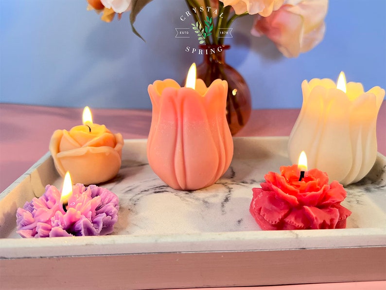Pode incluir: Uma cole&ccedil;&atilde;o de velas em forma de flor em v&aacute;rias cores, incluindo rosa, laranja e roxo, exibidas em uma bandeja de m&aacute;rmore. Cada vela tem um pavio aceso, criando um brilho quente. As velas s&atilde;o em forma de rosa, tulipa e outros desenhos florais.