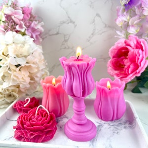 Pode incluir: Uma cole&ccedil;&atilde;o de velas em forma de flores em tons de rosa e f&uacute;csia, com pavios acesos. As velas est&atilde;o dispostas numa bandeja branca, rodeadas por flores artificiais. As velas incluem desenhos de tulipas e rosas.