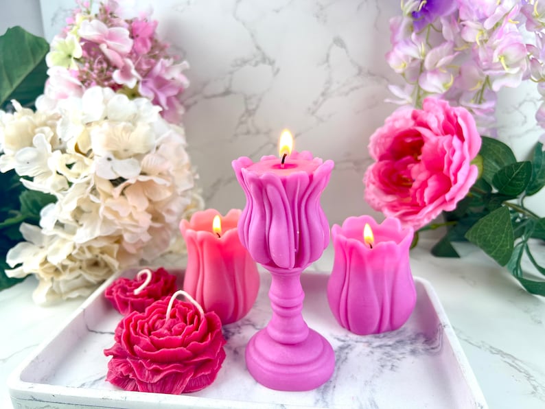 Pode incluir: Uma cole&ccedil;&atilde;o de velas em forma de flores em tons de rosa e f&uacute;csia, com pavios acesos. As velas est&atilde;o dispostas numa bandeja branca, rodeadas por flores artificiais. As velas incluem desenhos de tulipas e rosas.