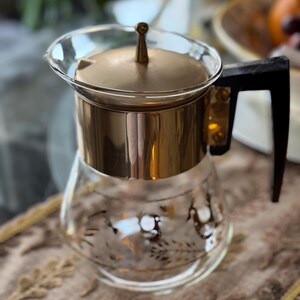 May include: A vintage glass and gold-toned coffee carafe with a black handle. The carafe has a clear glass body with a gold floral design and a gold lid. The carafe is sitting on a patterned surface.