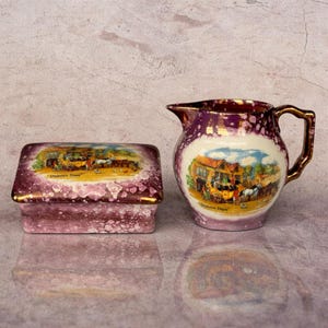 May include: A decorative ceramic pitcher and rectangular box set. The set features a purple and white speckled design with gold trim. Each piece has a central image of a horse-drawn carriage and the text "Dickens' Days".