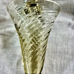 May include: A clear, spiral-textured glass vase with a flared top and a rounded base. The vase has a light amber tint and is designed with a twisted pattern. The glass reflects light, creating highlights and shadows.