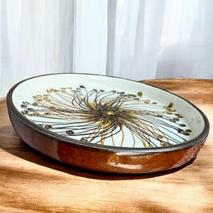 Può includere: Piatto ovale in ceramica con bordo e base marrone. L'interno presenta uno sfondo azzurro chiaro con un motivo floreale centrale marrone e giallo. Il piatto è posto su una superficie di legno.