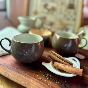 May include: A round, speckled brown ceramic mug with a curved handle sits on a wooden board next to a small white dish holding two cinnamon sticks. A second, similar brown mug and a blurred ceramic pitcher are visible in the background.