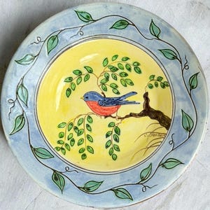 May include: A hand-painted ceramic plate featuring a blue and red bird perched on a brown branch with green leaves. The plate has a yellow center, a light blue border with green leaf accents, and a signature in the bottom right corner.