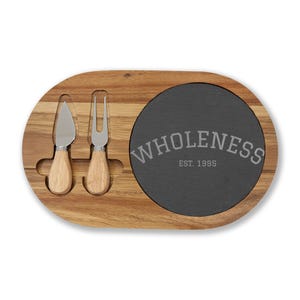 May include: Wooden cheese board with a dark gray slate insert. Includes a cheese knife and fork with wooden handles. The slate insert has the word "WHOLENESS" and "EST. 1995" printed on it.