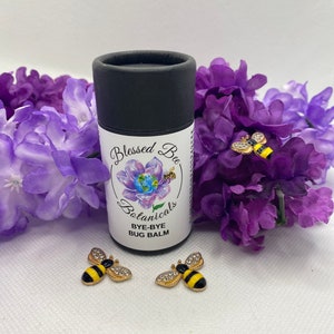 Puede incluir: Un tubo negro con una etiqueta blanca que dice "Blessed Bee Botanicals Bye-Bye Bug Balm". Dos figuritas de abejas doradas con rayas negras y amarillas y pedrería están sobre una superficie blanca.