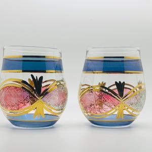 May include: Two clear, stemless glasses with blue bands and gold accents. The glasses feature pink and gold decorative elements. The design includes a black and gold floral motif. The glasses are set against a white background.