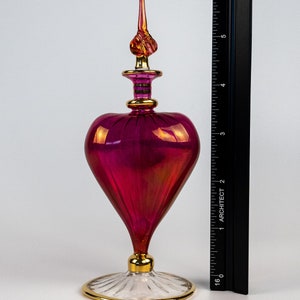 May include: A heart-shaped glass bottle with a gold-trimmed stopper and base. The bottle is a deep pink color with iridescent highlights.