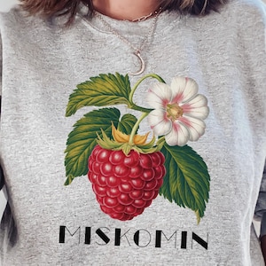 Op de afbeelding: Grijs sweatshirt met een afbeelding van een rode framboos en een witte bloem met groene bladeren. De tekst "MISKOMM" is onder de afbeelding gedrukt.