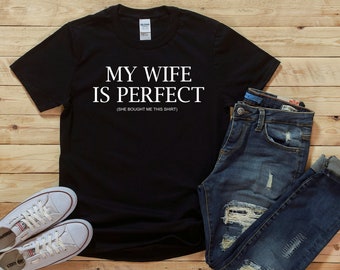 May include: Black t-shirt with white text that reads "My wife is perfect (She bought me this shirt)". The shirt is displayed on a wood background with a pair of white sneakers and blue jeans.