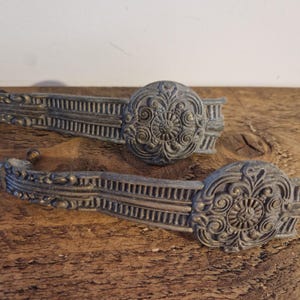 May include: A pair of antique bronze-effect drawer pulls. Each features an elaborate floral and scroll design, with a central circular motif and a curved handle with linear detailing. The hardware is displayed on a rustic wooden surface.