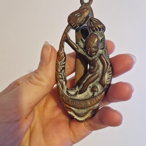May include: A brass door knocker featuring a detailed design of a fairy holding a mushroom. The knocker is oval shaped and has a patina finish.