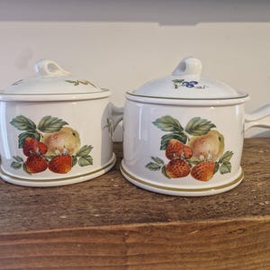 Può includere: Due pentole bianche in ceramica con coperchi e manici, decorate con un motivo a fragole e pere. Le pentole hanno una forma arrotondata e poggiano su una superficie di legno. I coperchi hanno una piccola maniglia ad anello.