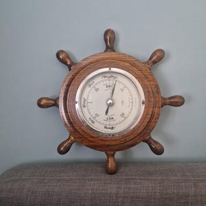 May include: A wooden ship's wheel barometer with a silver-colored dial. The dial has a needle pointing to the current atmospheric pressure, with text indicating "Very Dry" and "Wet". The wheel has eight spokes and is mounted on a gray surface.