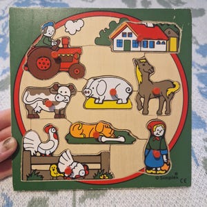 Può includere: Puzzle in legno con animali della fattoria, con bordo verde e design circolare rosso. Il puzzle presenta un trattore, una casa, un cavallo, una mucca, un maiale, un cane, un gallo, una gallina e una ragazza. I pezzi hanno bottoni rossi.