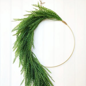 May include: A decorative wreath featuring a gold metal ring adorned with lush, vibrant green faux fern foliage. The fern cascades down the ring, creating a natural, elegant aesthetic. The wreath is suspended by a natural twine loop, perfect for home decor.