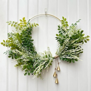May include: A decorative wreath featuring a gold metal ring base adorned with lush green foliage and delicate white accents. The wreath is embellished with three small, gold-colored bells hanging from a twine bundle. The wreath is hanging on a white, vertical-paneled wall.