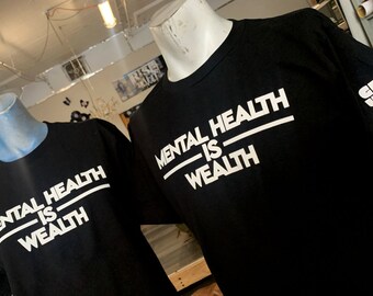 Puede incluir: Dos camisetas negras con texto blanco que dice "Mental Health Is Wealth" en una fuente gráfica y en negrita. Las camisetas se muestran en maniquíes.