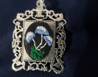 May include: A silver framed pendant necklace with an embroidered image of three white and blue mushrooms on a black background. The mushrooms have green stems.