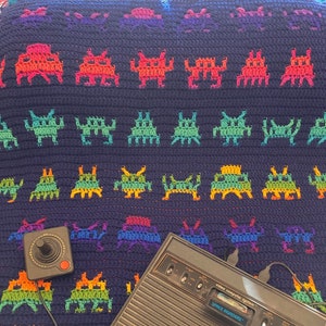 May include: A vintage Atari video game console with a joystick and a crocheted blanket featuring a repeating pattern of colorful space invaders. The console has a label that reads "video computer system" and "space invaders".